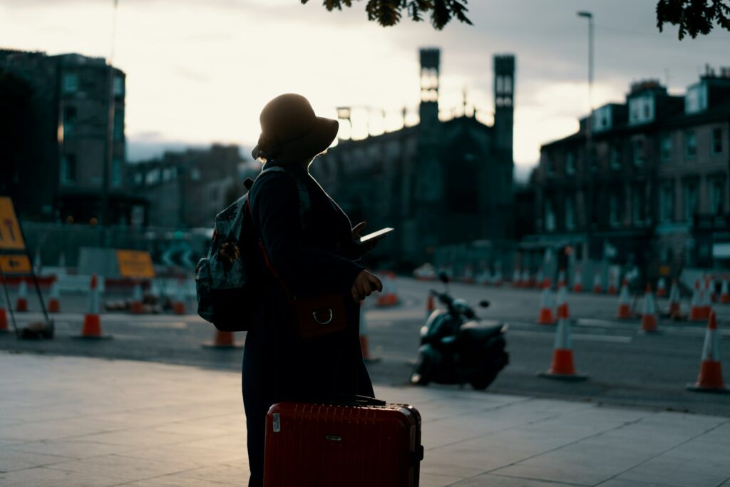 woman wearing coat standing on road with travel luggage during daytime thinking Hostel Vs Hotel Which Option Suits Best?