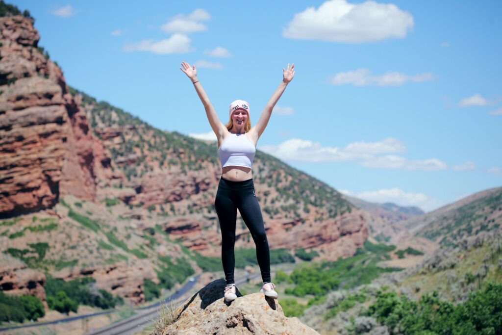 a woman standing on a rock with her arms raised happy because she just read Unconventional Ways to Save Money While Traveling